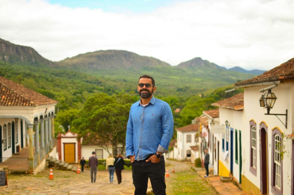 Man posing on a cobblestone street in a picturesque village with mountains in the background.